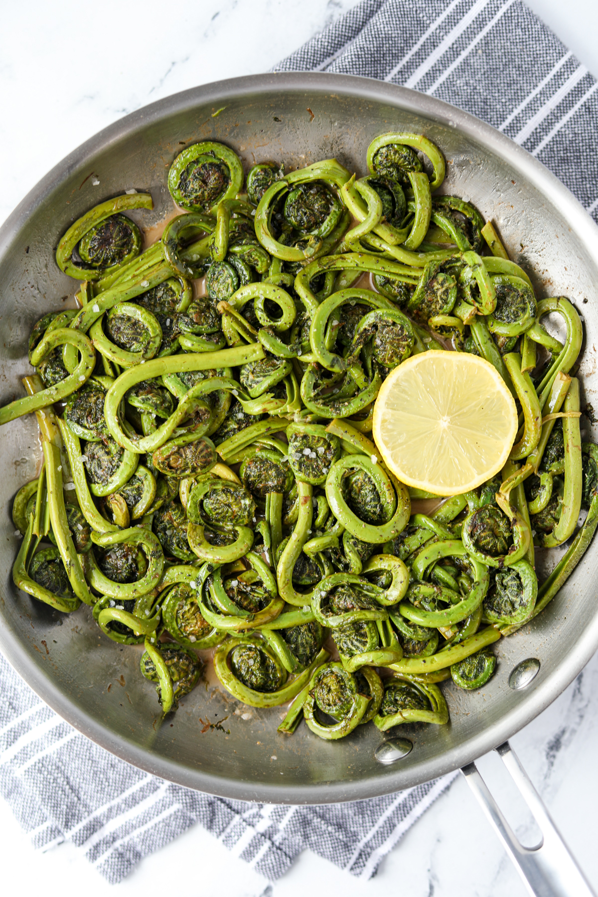 A skillet with panfried fiddleheads, garnished with a slice of lemon.