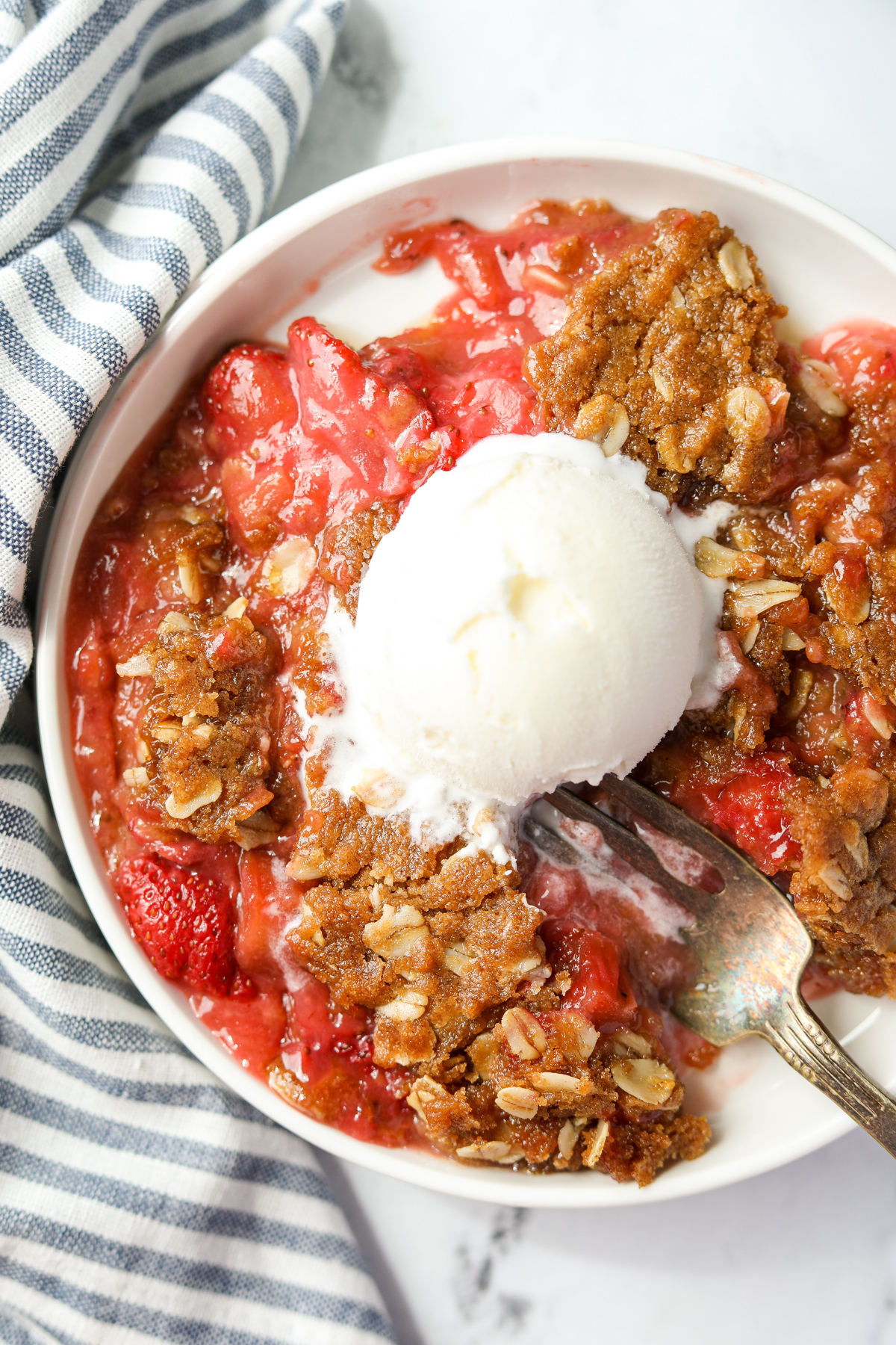 A bowl of rhubarb crisp with strawberries, and served with a scoop of vanilla ice cream.