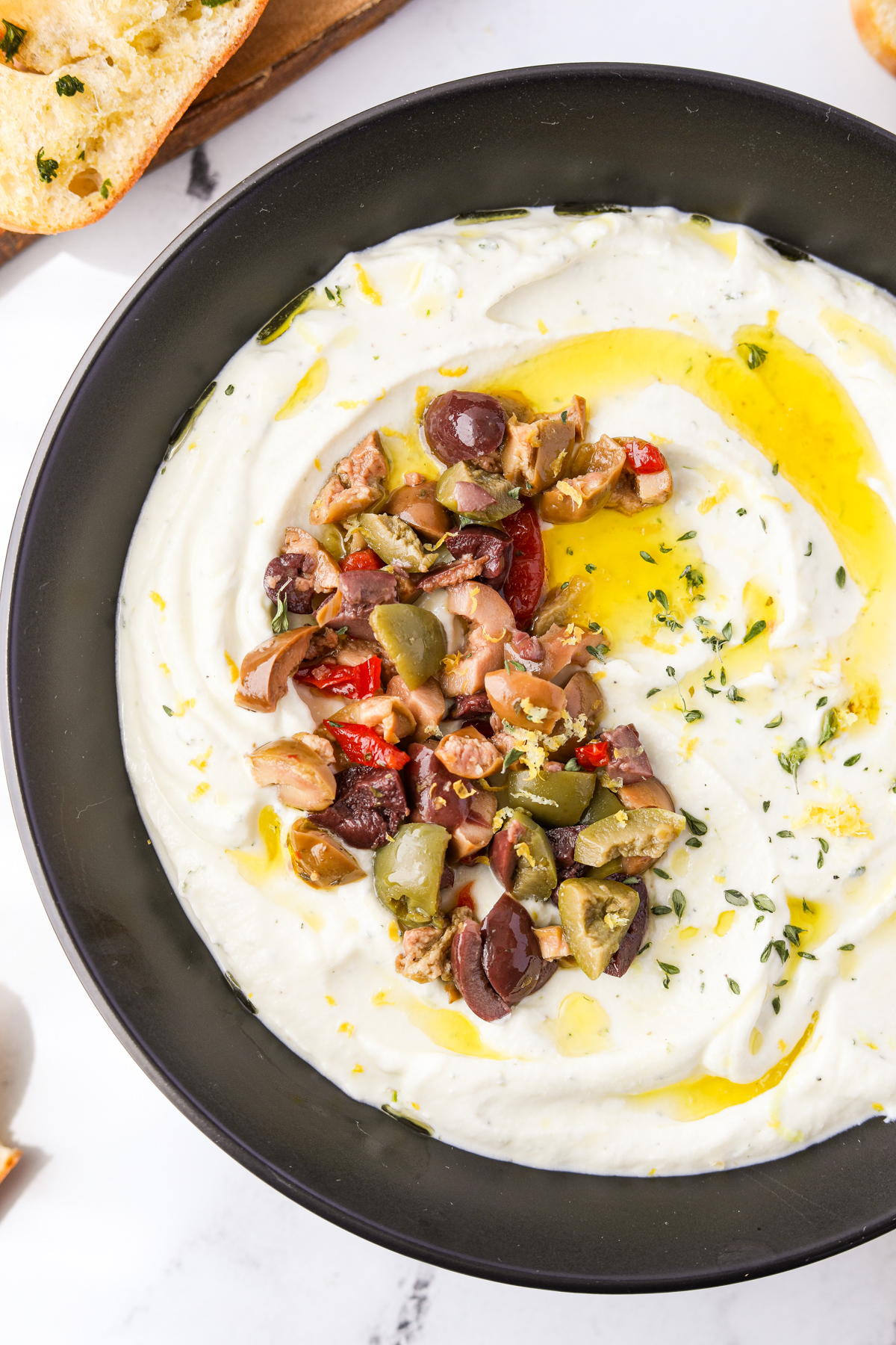 A bowl with a cheesy dip inside, garnished with greek olives and olive oil.