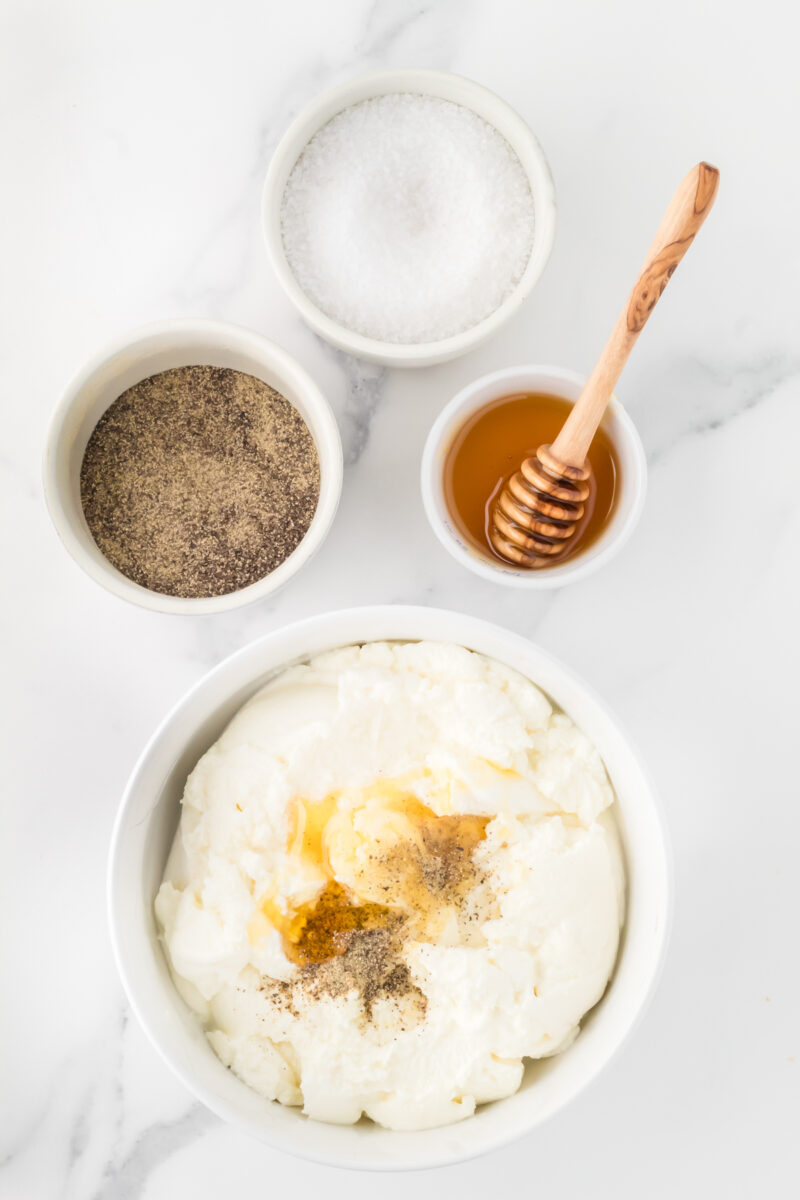 A bowl filled with ricotta, surrounded by smaller bowls containing honey, salt, and pepper.