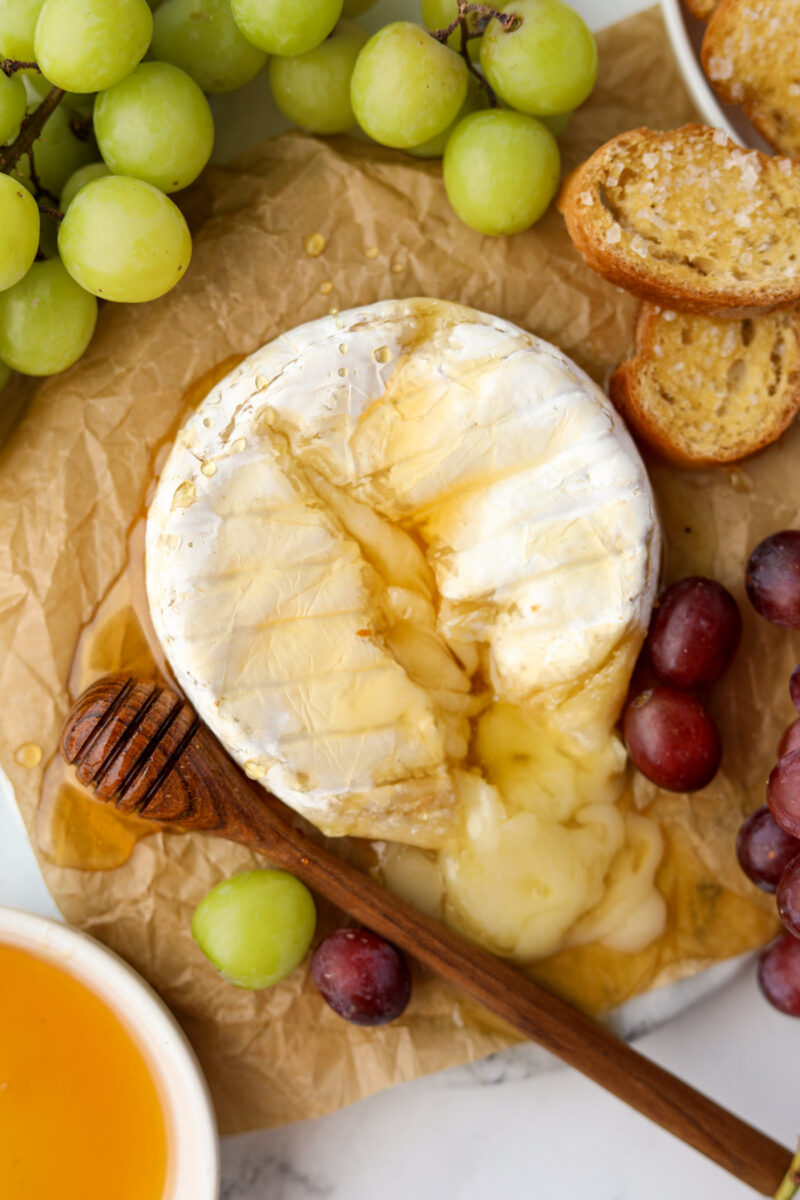A wheel of melty brie surrounded by grapes, honey, and toasted baguette slices.