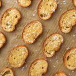Toasted baguette slices on a parchment lined baking sheet, topped with flaky salt.